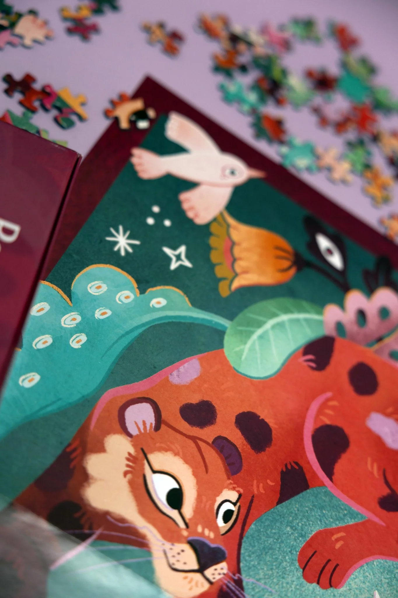 Primer plano de la página colorida de un libro infantil ilustrado con un jaguar tumbado sobre hojas verdes, con un fondo de plantas y patrones caprichosos, junto a piezas de puzzle dispersas al fondo.