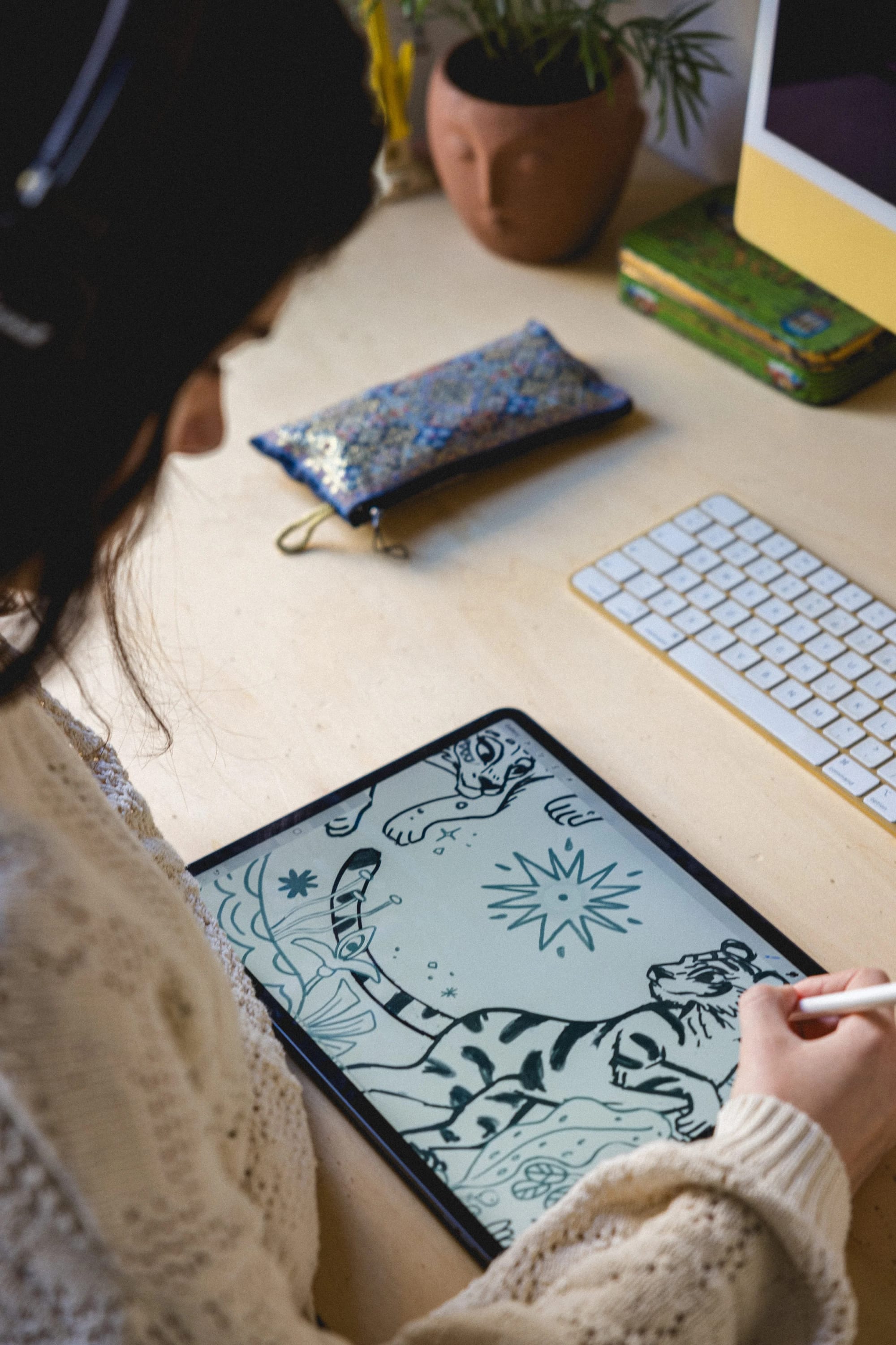 Una persona dibujando un boceto en blanco y negro de un tigre en una tableta, sentada en un escritorio con un teclado, un bolso colorido y objetos decorativos como una planta y una maceta con forma de cara.