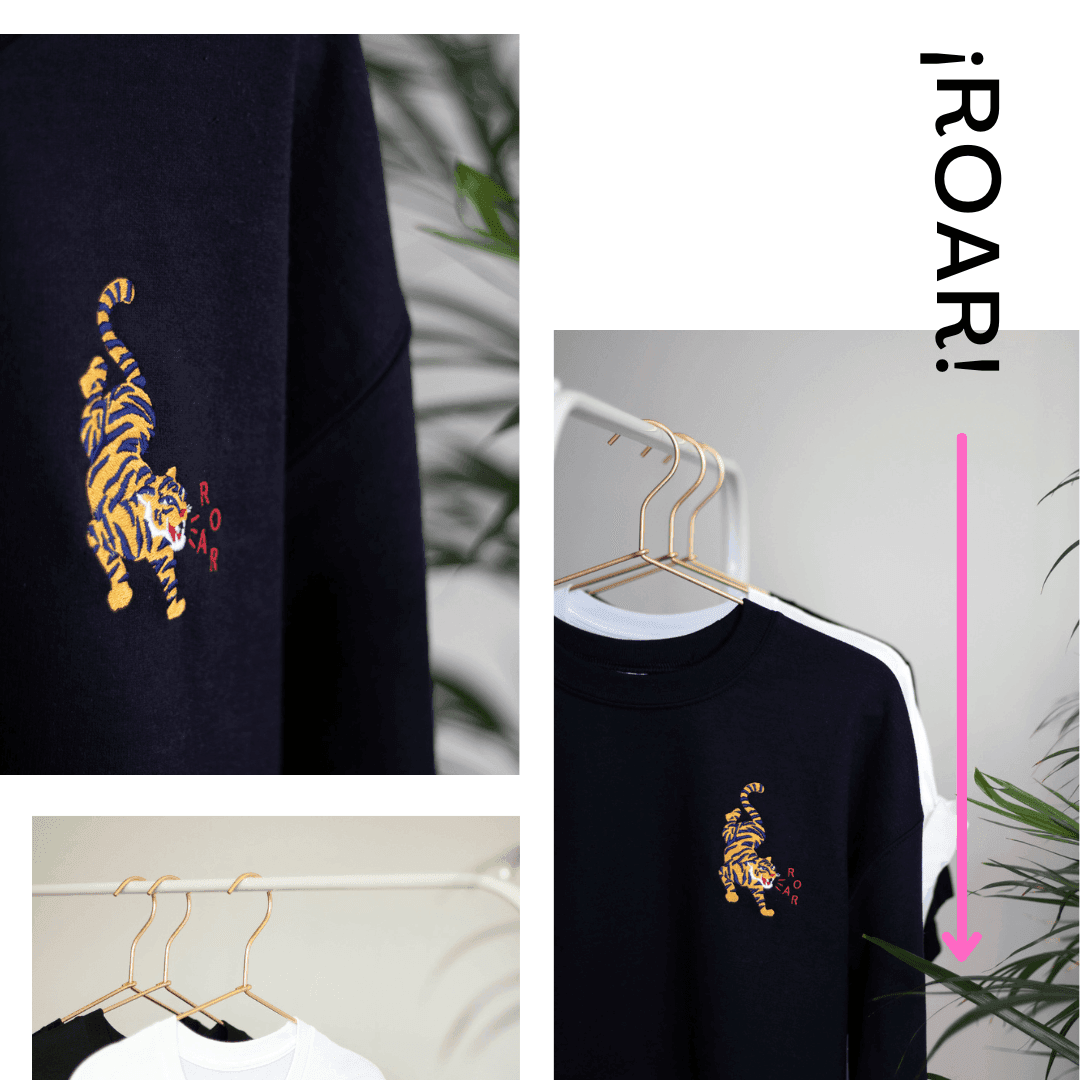Nahaufnahme eines marineblauen Sweatshirts mit einem gelb-schwarz bestickten Tiger und dem Wort 'ROAR' auf einem Kleiderbügel, mit weiteren Sweatshirts daneben und grünen Pflanzen im Hintergrund.