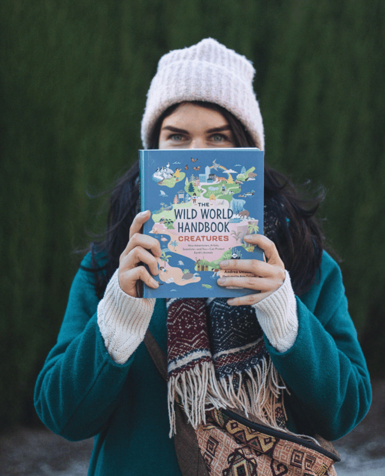 Eine Frau mit rosa Strickmütze und türkisem Mantel hält ein Buch mit dem Titel 'The Wild World Handbook: Creatures' vor ihr Gesicht und steht draußen vor Bäumen.