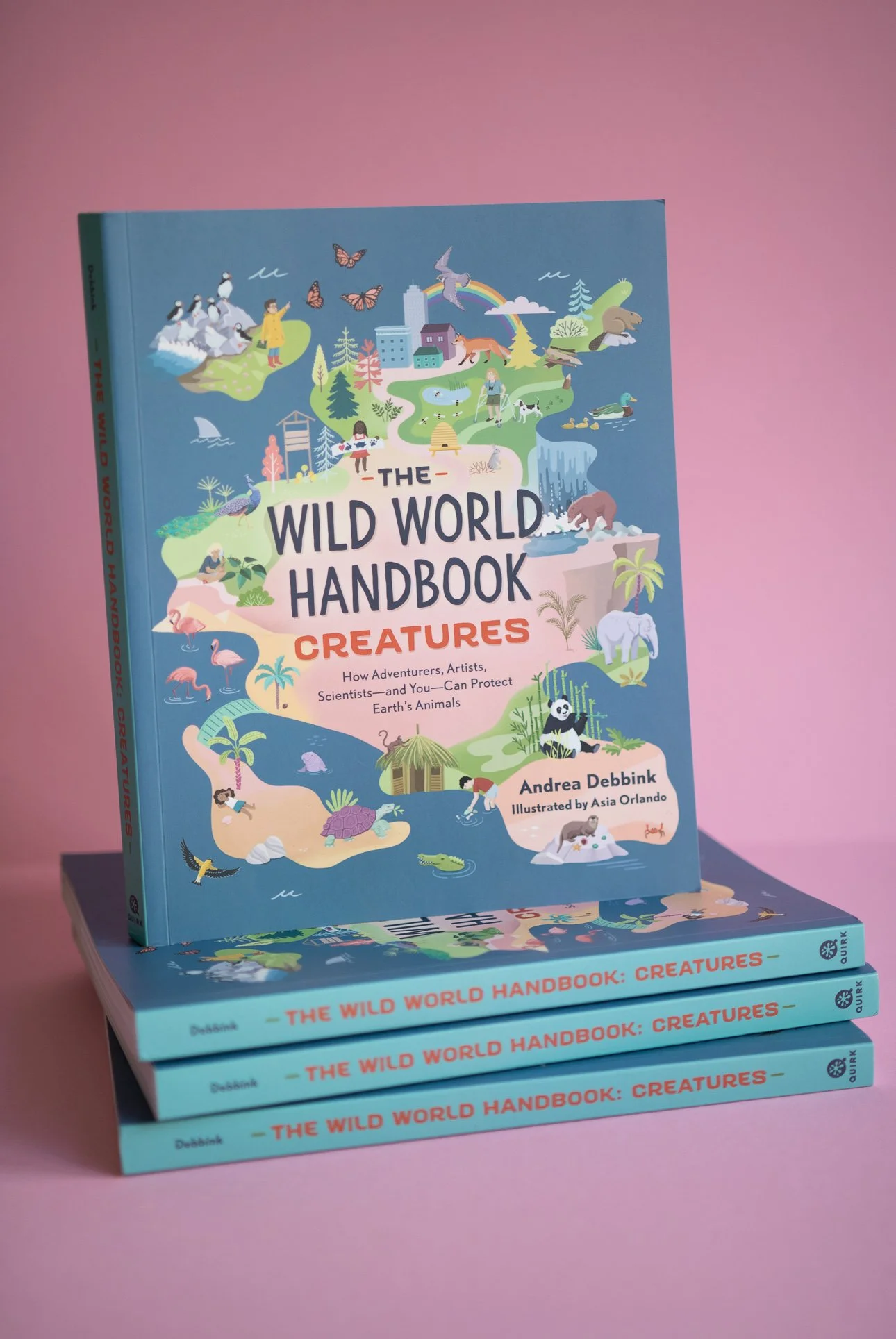 Ein farbenfrohes Kinder-Hardcoverbuch mit dem Titel "The Wild World Handbook: Creatures" steht aufrecht auf einer rosa Fläche, weitere Exemplare sind davor gestapelt. Das Cover zeigt Illustrationen verschiedener Tiere und Naturszenen.