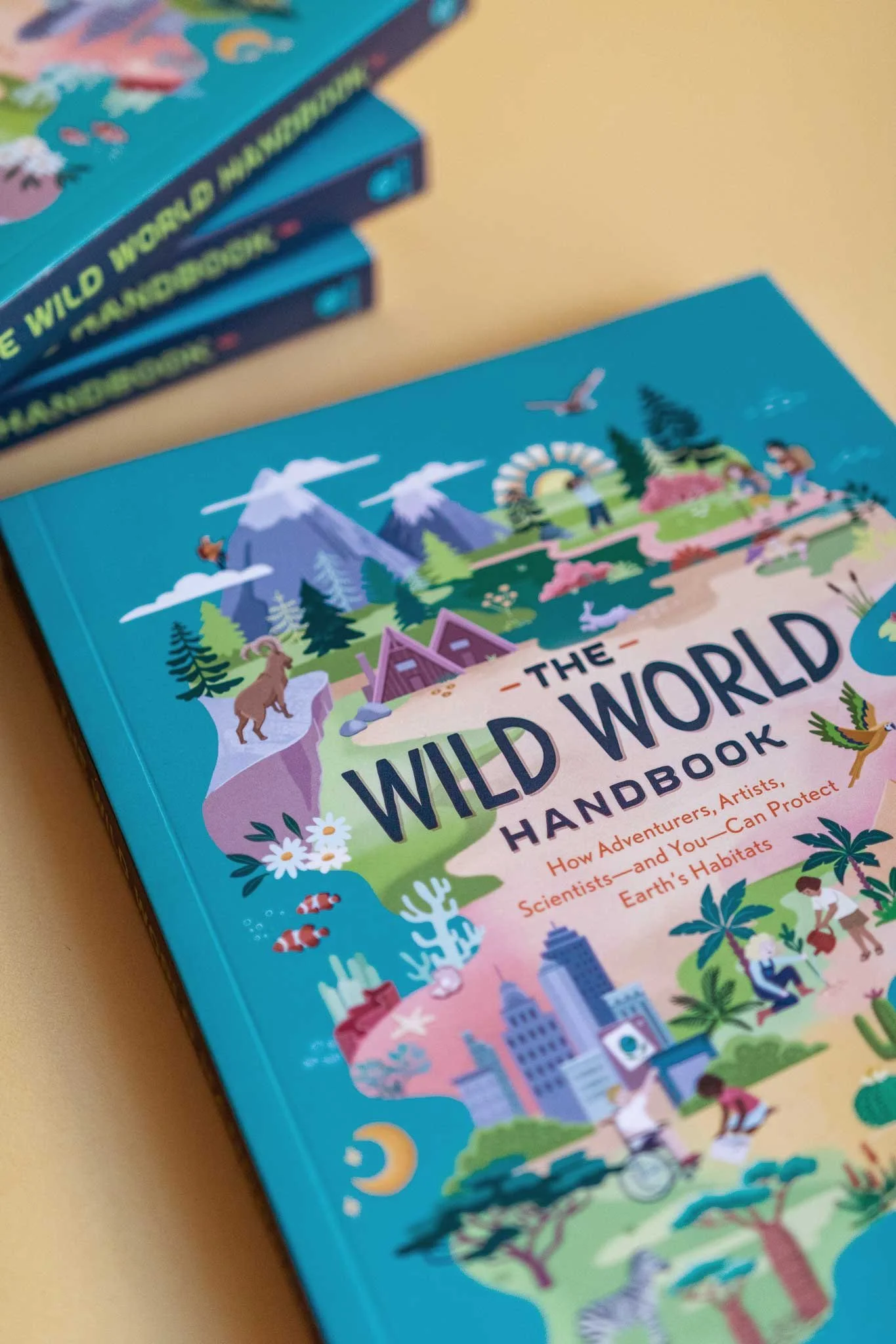Das Cover eines Kinderbuchs mit dem Titel 'The Wild World Handbook' zeigt farbenfrohe Illustrationen von Bergen, Wäldern, Tieren und Menschen bei Outdoor-Aktivitäten.