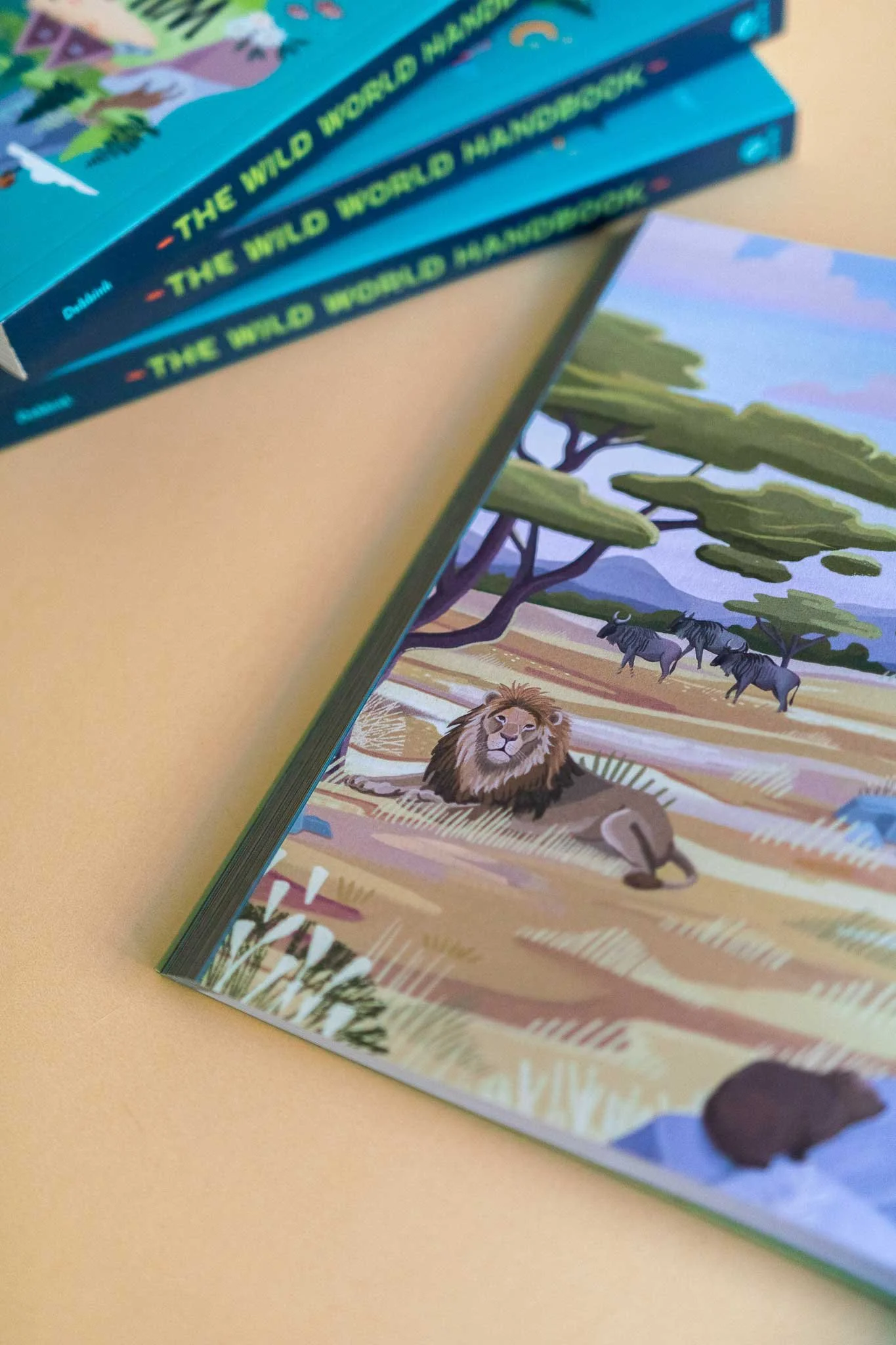 Aufgeschlagenes Kinderbuch mit einer illustrierten Safari-Szene mit einem Löwen, Zebras und einem Nilpferd, mit weiteren Büchern daneben gestapelt.