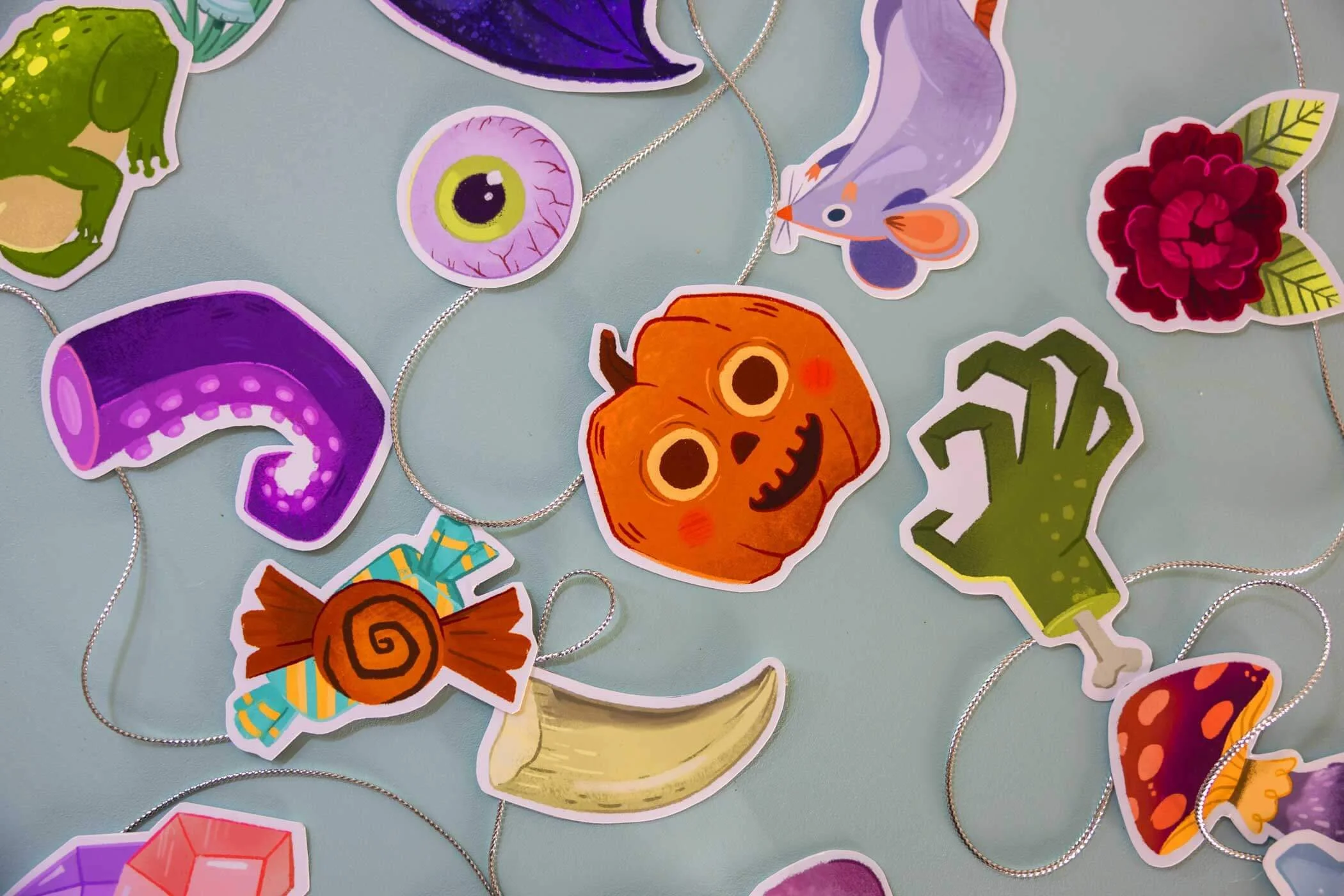 Pegatinas de papel y bisutería con temática de Halloween: una calabaza, un tentáculo morado, un ojo, una rana, una mariposa, una flor, una seta, una mano, un plátano, un caramelo y otros elementos decorativos sobre un fondo claro.
