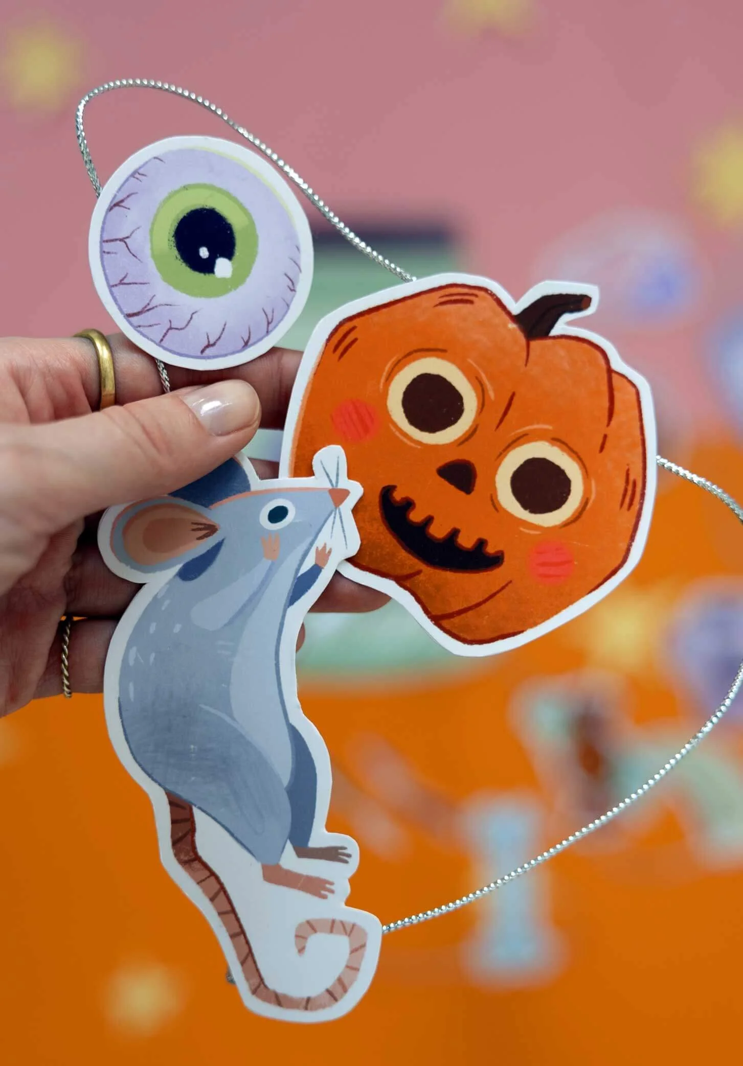 Mano sosteniendo tres recortes de papel temáticos de Halloween: un ojo, una calabaza tallada y un ratón gris, unidos por una cuerda, sobre un fondo naranja con decoraciones desenfocadas.