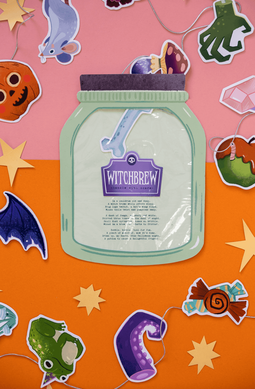 Composición temática de Halloween con una decoración en forma de tarro etiquetada como 'Witchbrew' y pegatinas variadas de Halloween: una calabaza, un murciélago, una lagartija, una araña, caramelos, luna y estrellas, sobre un fondo naranja y rosa.