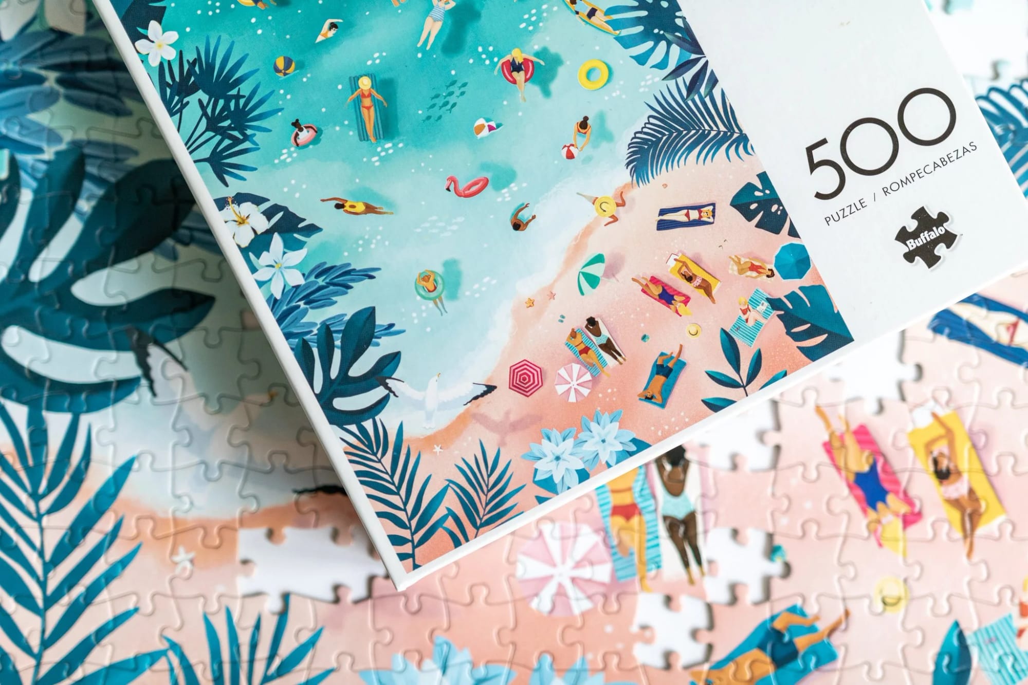 Une boîte de puzzle présentant une illustration colorée d’une scène de plage avec des personnes qui nagent, prennent le soleil et se détendent sur le sable, entourées d’un feuillage tropical et de fleurs.