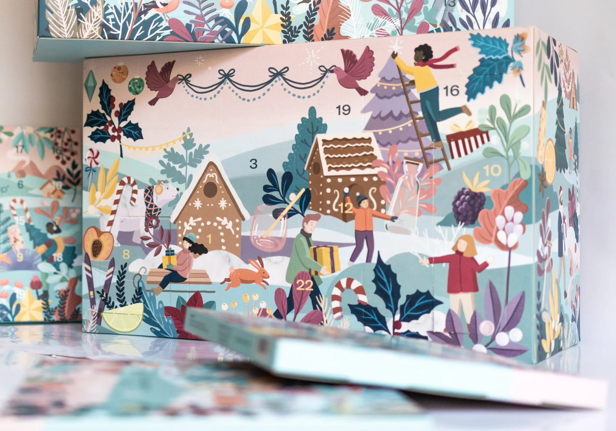 Illustrierte Weihnachtsszene auf einer Adventskalender-Schachtel mit Kindern, die einen Baum schmücken, Geschenke einpacken und in einer Schneelandschaft mit Lebkuchenhäusern, Stechpalme und Beeren spielen.