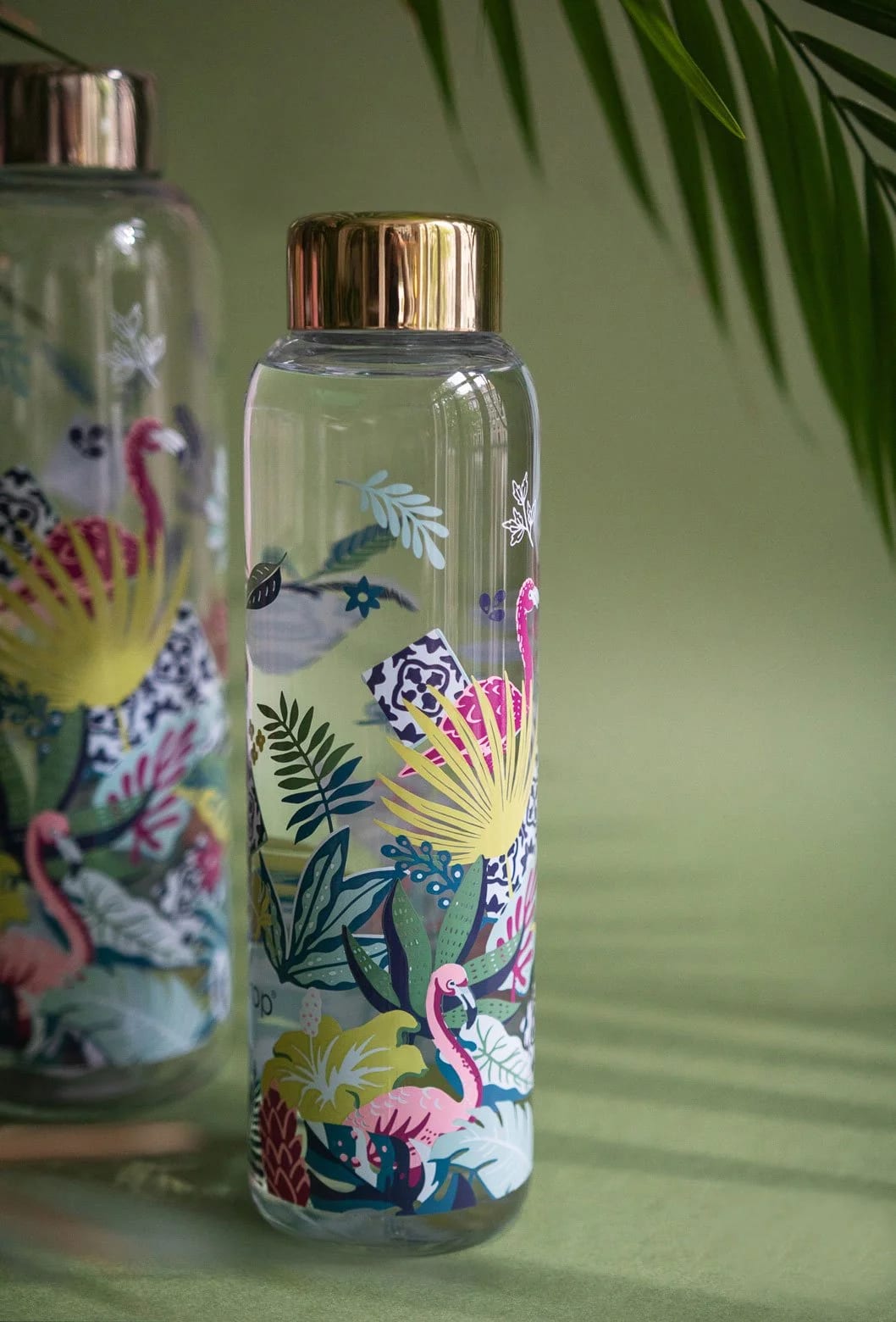Eine transparente Glas-Wasserflasche mit goldfarbenem Deckel, mit farbenfrohen tropischen Flamingo- und Blattmustern, auf einer grünen Oberfläche mit grünen Pflanzen im Hintergrund.