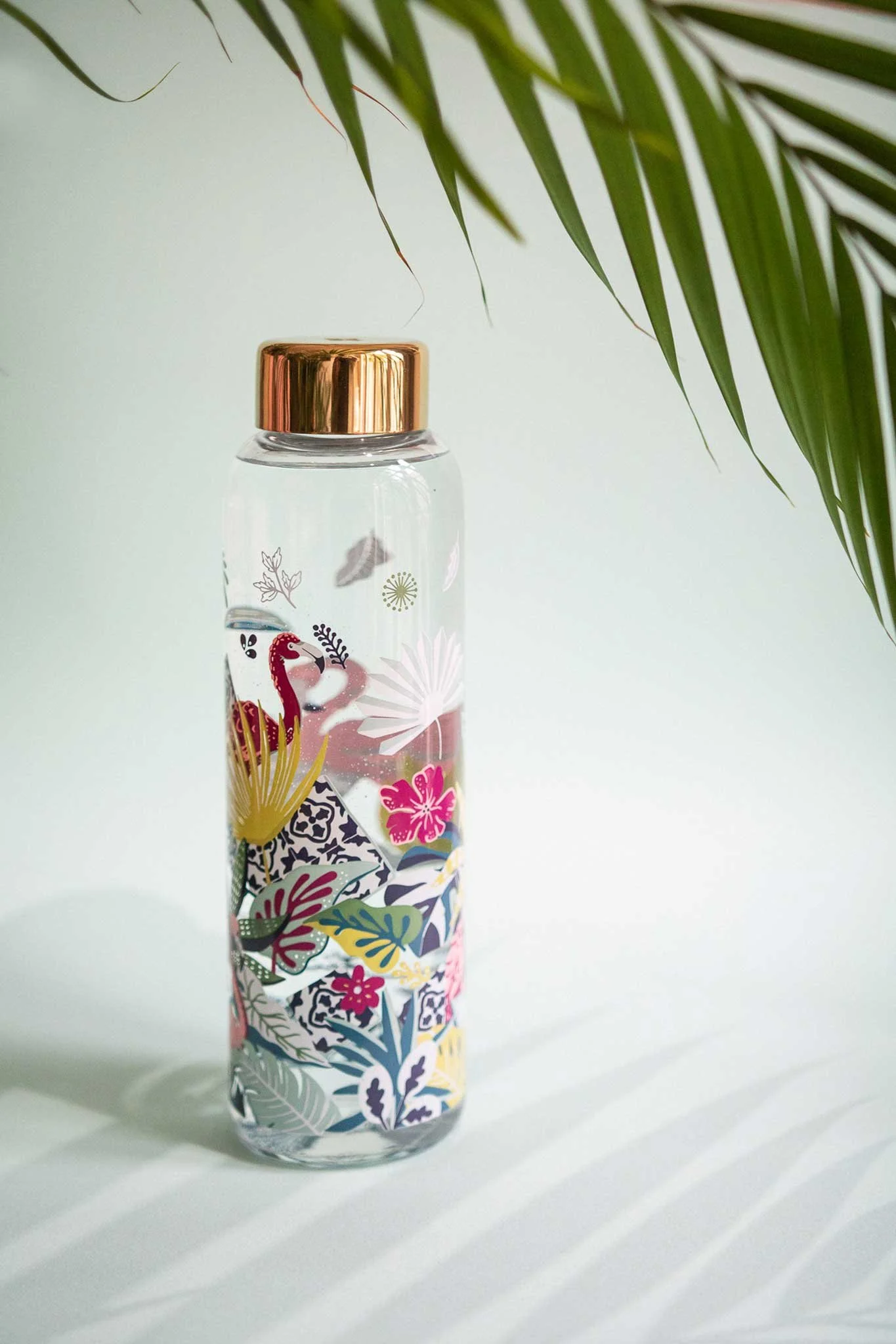 Eine transparente Wasserflasche mit goldener Kappe, dekoriert mit farbenfrohen tropischen Illustrationen, darunter ein rosa Flamingo, verschiedene Blätter und Blumen, vor schlichtem Hintergrund neben grünen Palmblättern platziert.