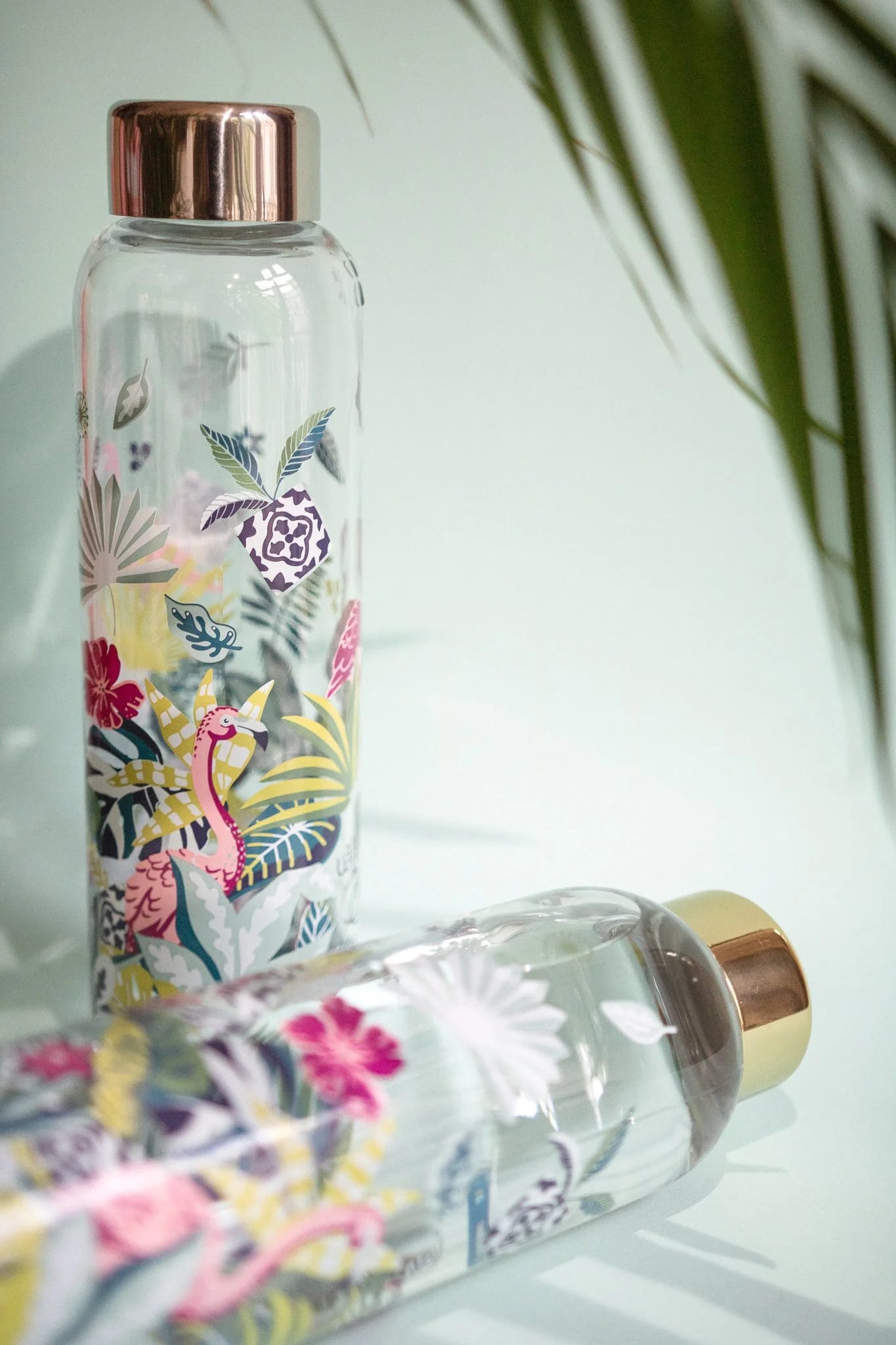 Zwei transparente Glas-Wasserflaschen mit farbenfrohen tropischen Blumen- und Dinosaurier-Illustrationen, eine steht aufrecht mit rosa Metallic-Kappe, die andere liegt auf der Seite mit goldener Metallic-Kappe. Oben rechts sind teilweise grüne Blätter sichtbar.